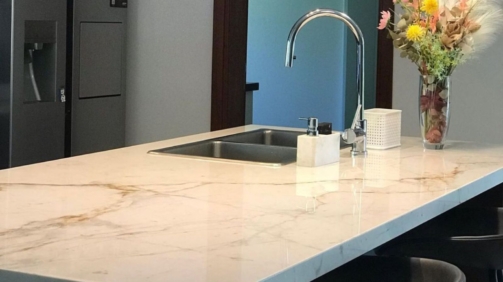 Foto de una mesada de cocina de cuarzo blanco simil mármol carrara. La cocina es moderna, tiene una bacha plateada y un jarrón con flores encima de la mesada.
